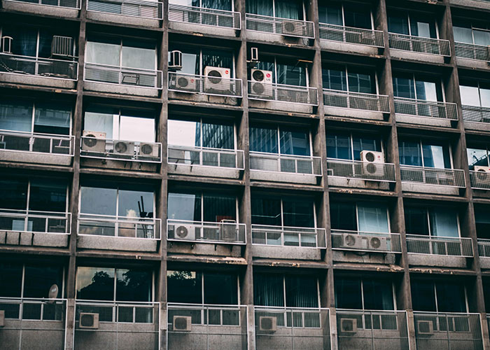 Grijs appartementsgebouw met tientallen energieverslindende airco-units op bijna elk balkon, wat wijst op hoge energieconsumptie en weinig duurzame koeling. De gevel oogt verouderd en uniform, met herhalende betonnen structuren en glazen schuifdeuren.
