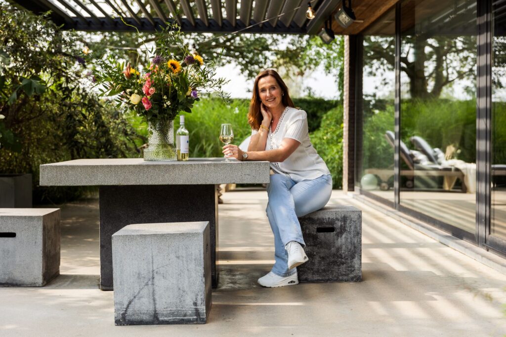 Vrouw zit ontspannen aan een betonnen tuintafel met bloemen en een glas wijn, onder een moderne terrasoverkapping met lamellen. De lamellen zorgen voor schaduw op het terras naast een woning met glazen schuifdeuren en uitzicht op een groene tuin.