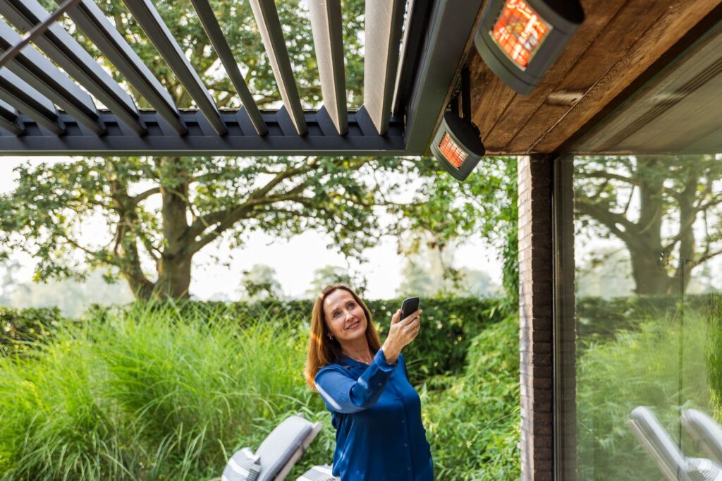 Vrouw bedient met een afstandsbediening de terrasverwarming onder een moderne terrasoverkapping met lamellen, bevestigd aan een woning met houten plafondaccenten. De lamellen zorgen voor schaduw en ventilatie, terwijl het buiten comfortabel blijft dankzij de warmtebron.