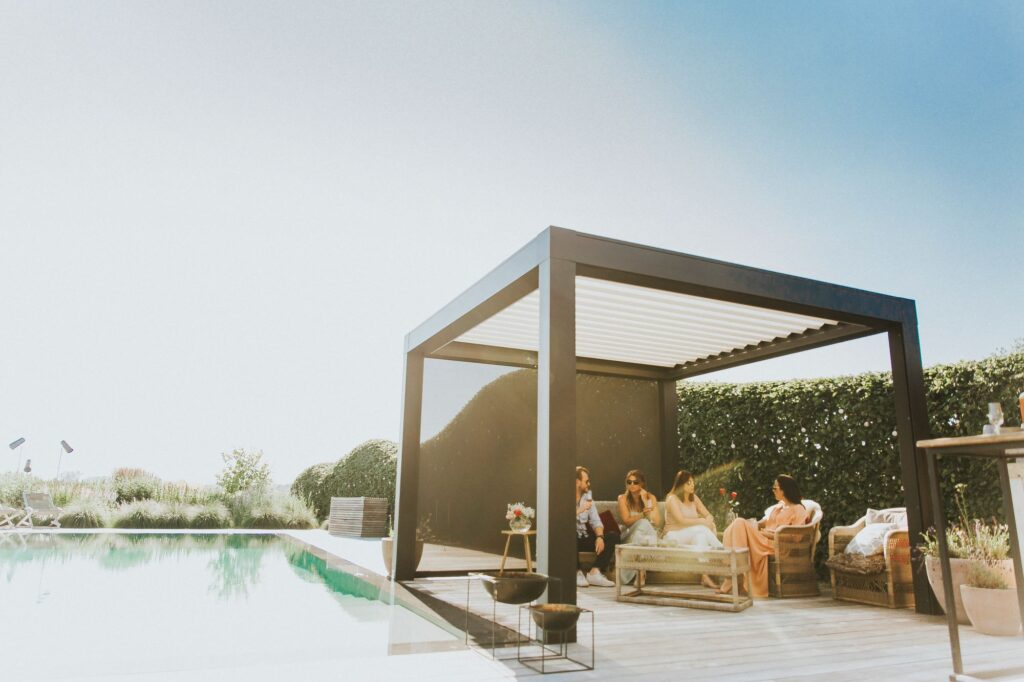 Vier mensen relaxen onder een moderne, open pergola naast een zwembad, omringd door groene hagen en zonnig weer. Ze zitten op comfortabele loungebanken en praten gezellig met elkaar in een stijlvolle buitenruimte.